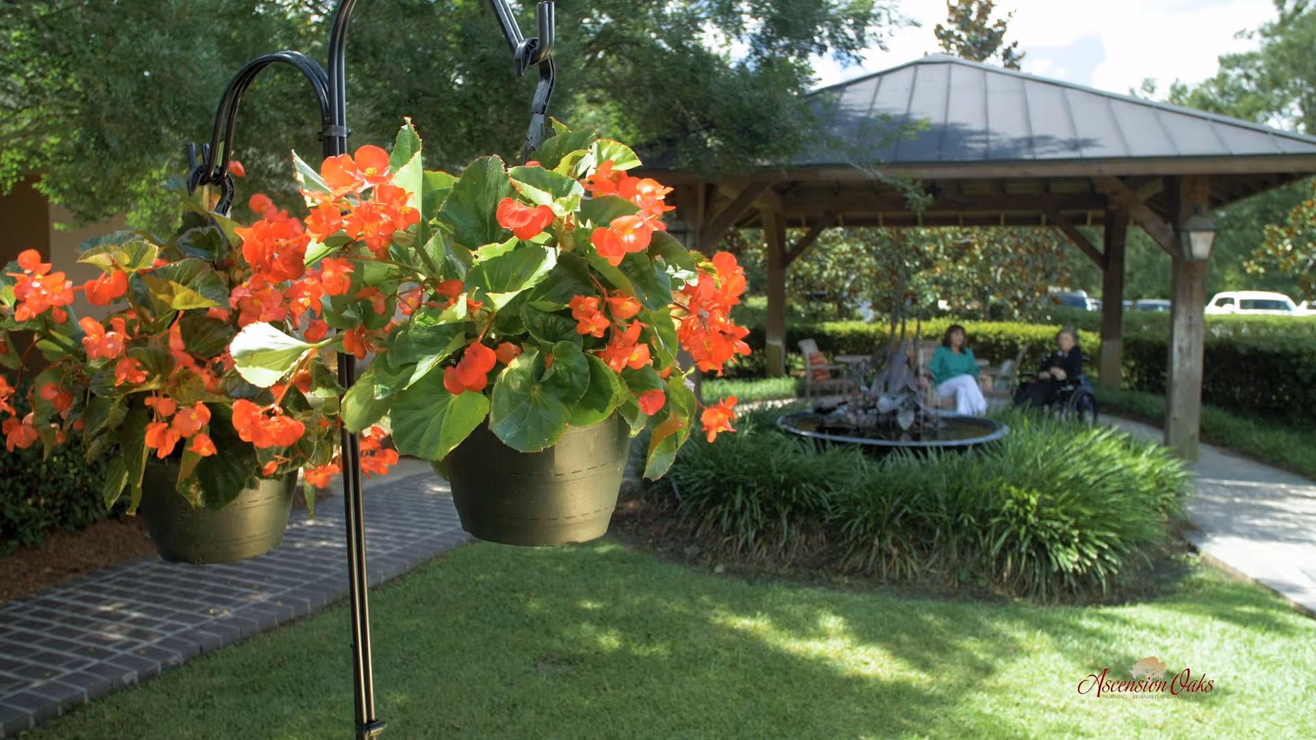 Two hanging pots filled with vibrant orange flowers in the foreground, with a gazebo and two people sitting on chairs near a small fountain surrounded by greenery in the background.