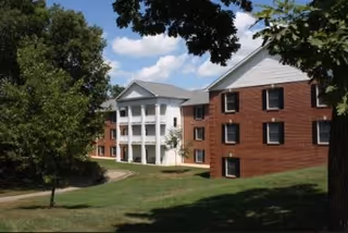 Exterior view of a multi-story brick building with white columns and a sloped roof, surrounded by green grass and trees under a partly cloudy sky.