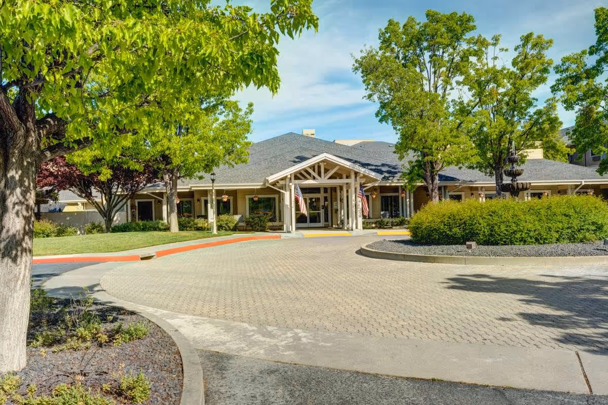 Front exterior view of The Village at Rancho Solano facility showing a circular driveway with a landscaped roundabout, green trees, and a building entrance with a covered porch and American flags.