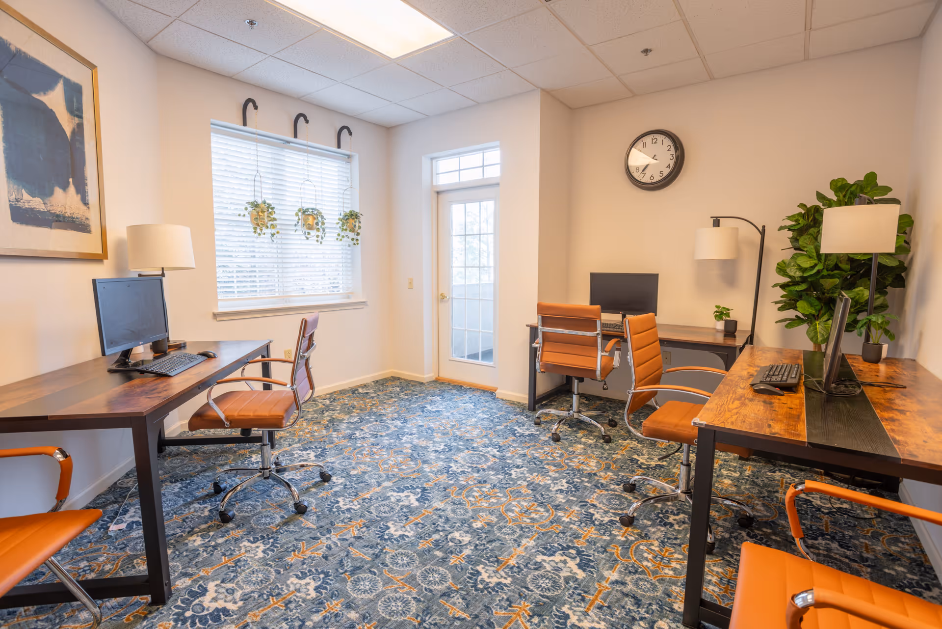 A bright office room with three wooden desks, each equipped with a computer monitor and keyboard. There are four orange swivel chairs around the desks. The room has a blue patterned carpet, a large window with three hanging plants, a door with glass panels, a wall clock, two floor lamps, a potted plant in the corner, and a framed abstract painting on the wall.