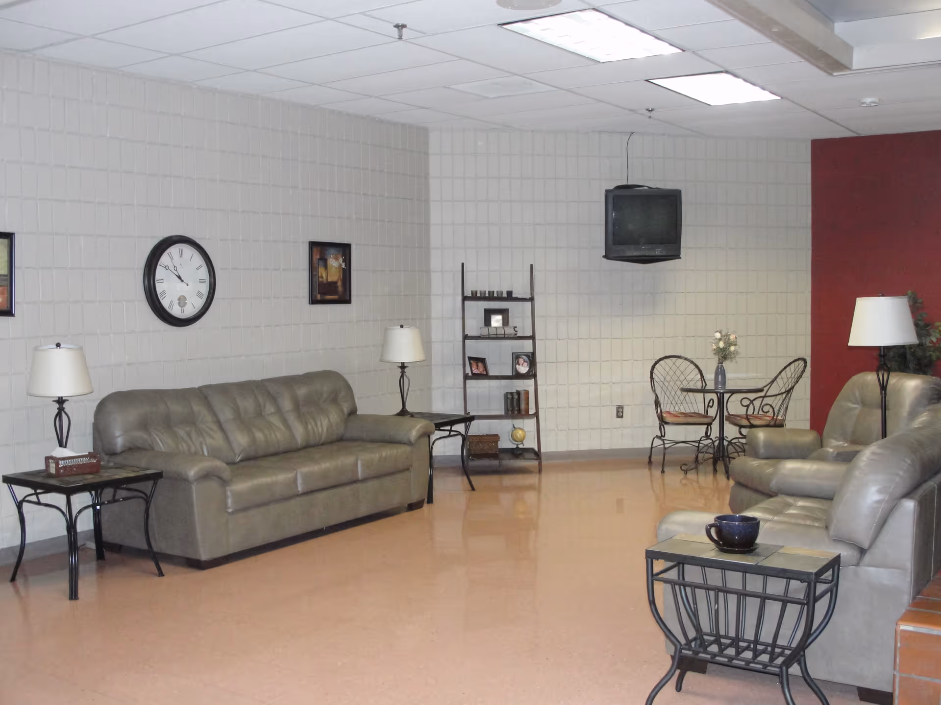 A senior living facility common area with beige leather sofas and armchairs, two side tables with lamps, a wall clock, framed pictures on the wall, a small round table with two wrought iron chairs, and a wall-mounted television. The walls are light-colored with one red accent wall.