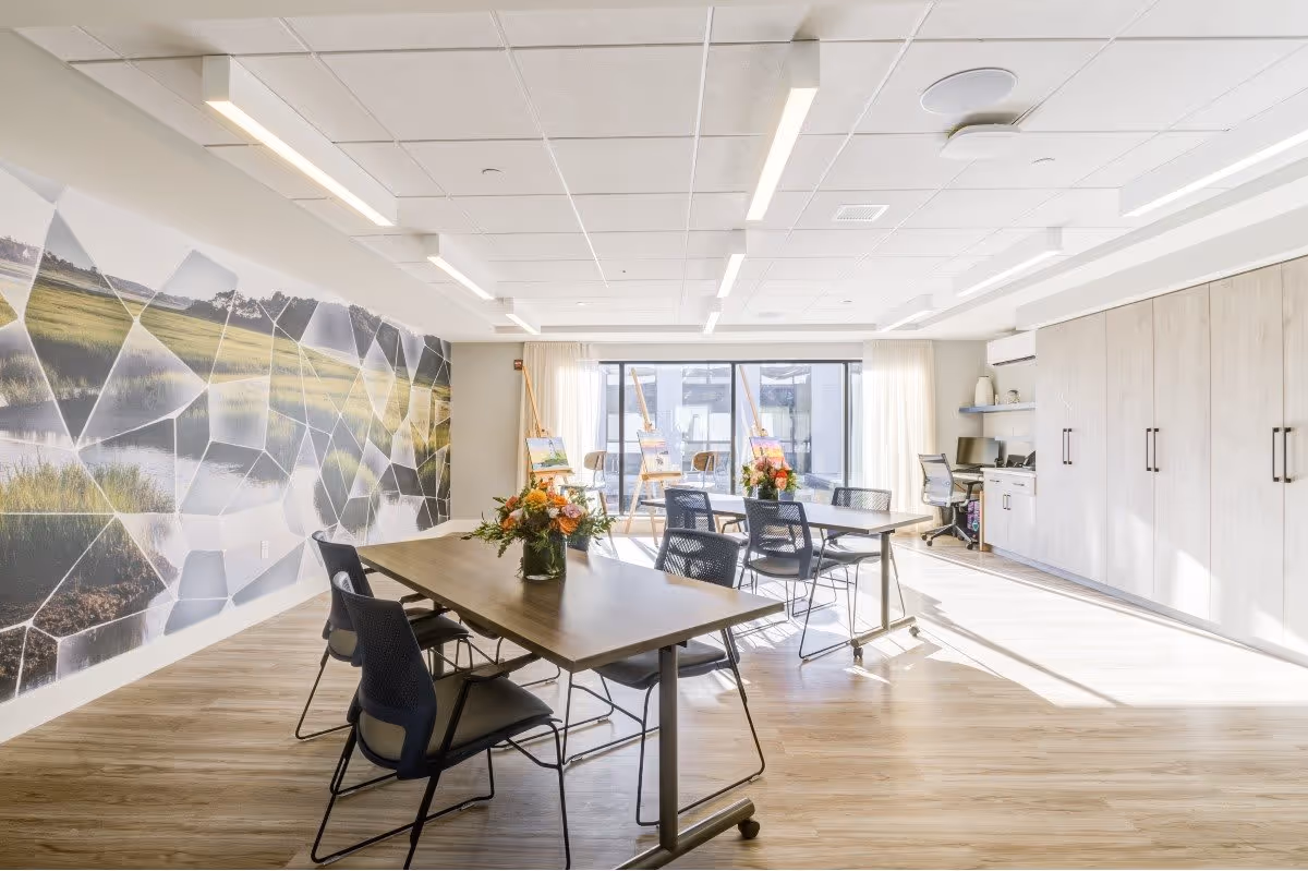 A bright, modern activity room with wooden floors and large windows letting in natural light. The room features two tables with black chairs, flower arrangements on the tables, a wall mural depicting a nature scene, and easels with paintings near the window. There is also a built-in desk with a computer and storage cabinets along one wall.