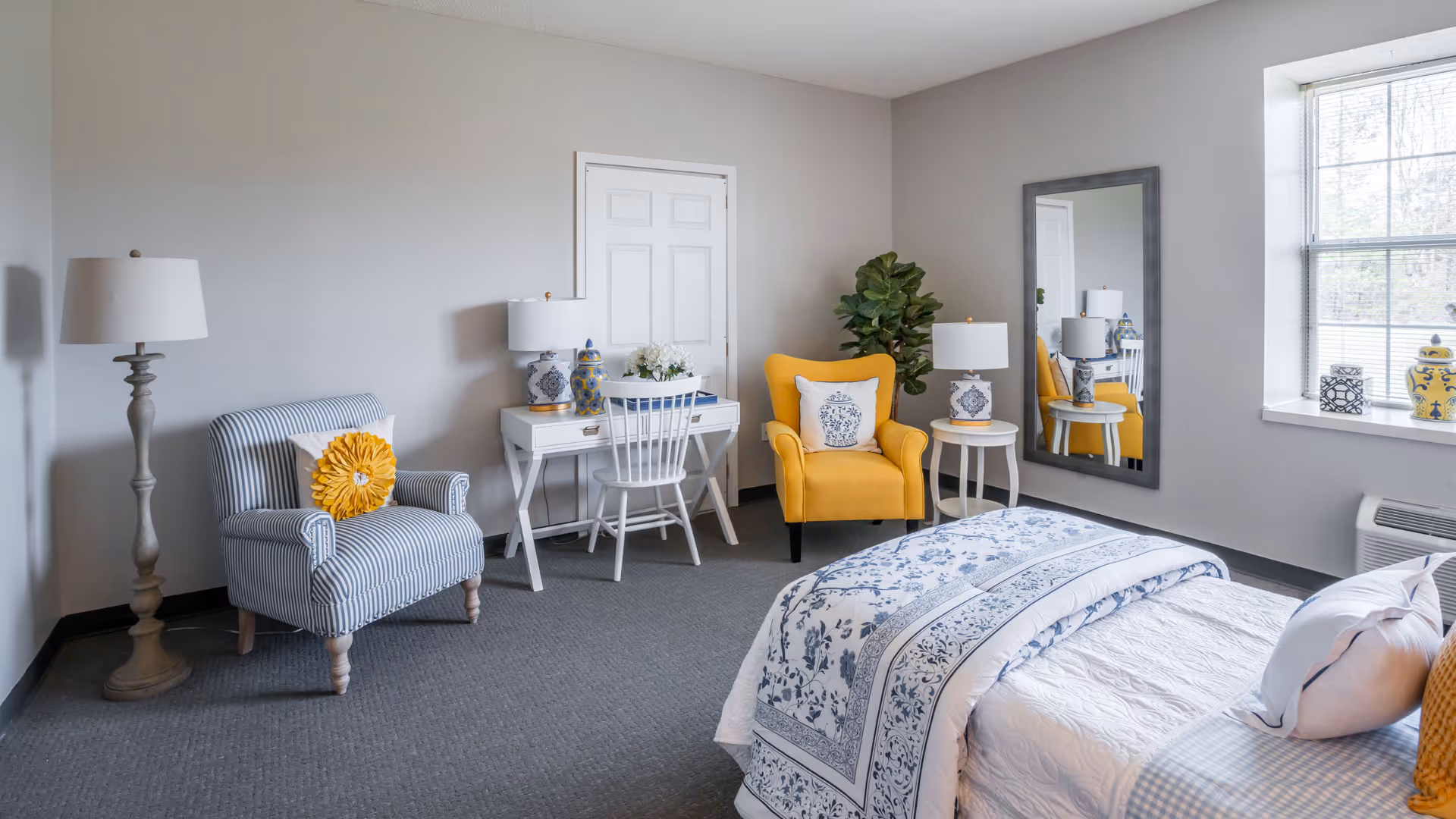 Bright furnished bedroom with a bed, blue-and-white bedding, a yellow accent chair, striped armchair, desk, mirror, and window.