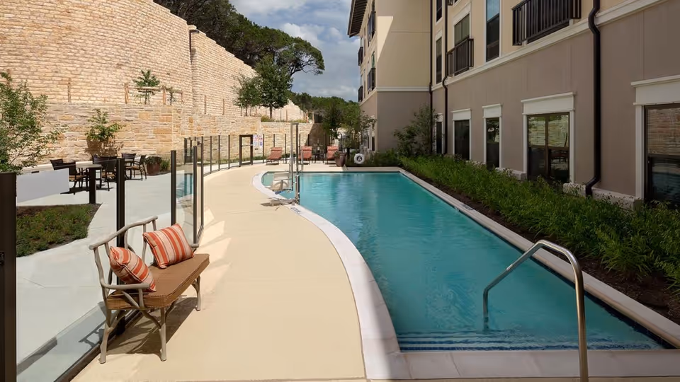 Outdoor swimming pool area at Belmont Village Senior Living West Lake Hills with a narrow pool, a bench with striped cushions, glass fencing, patio tables and chairs, and a stone wall in the background under a partly cloudy sky.