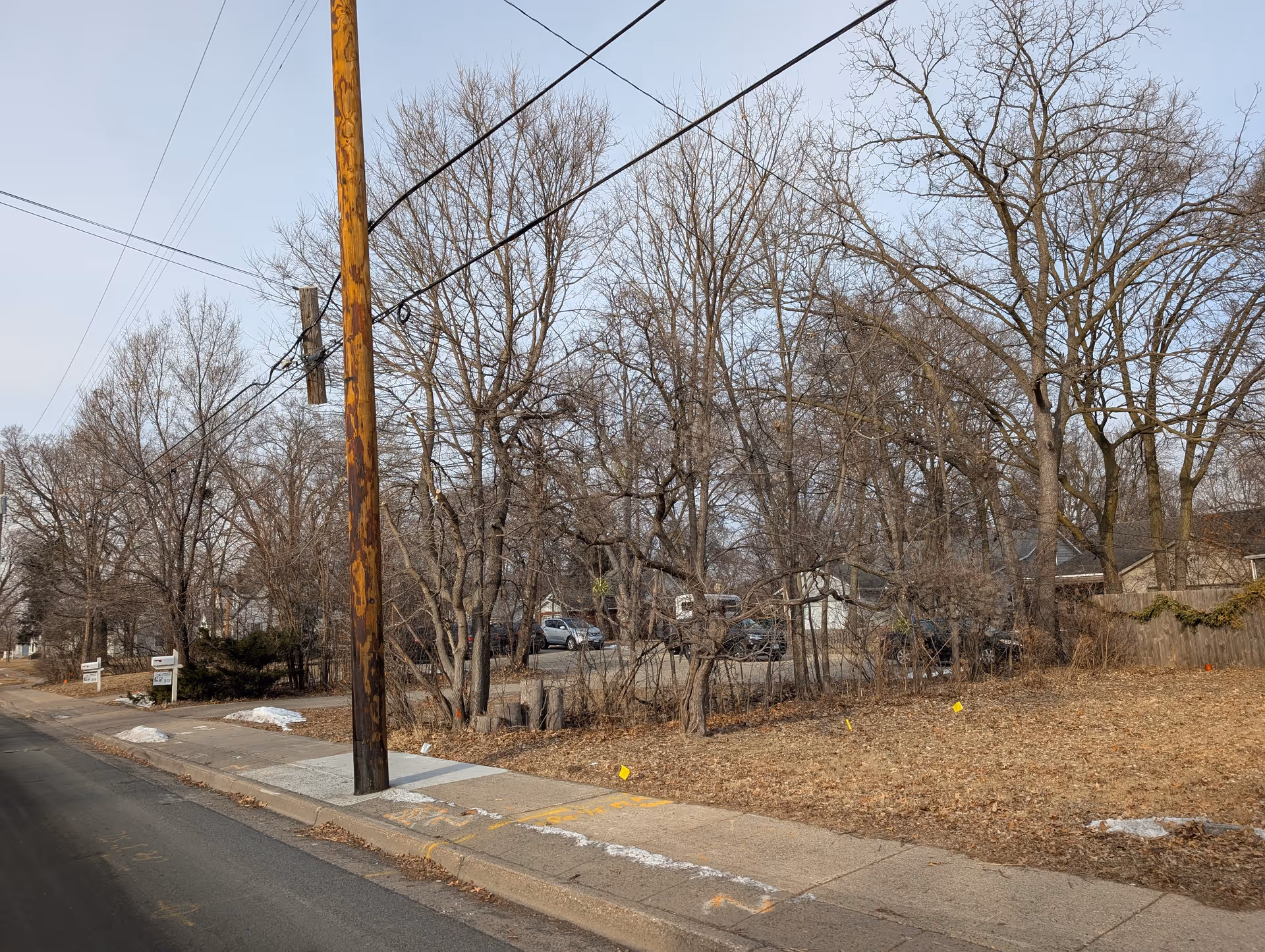A street view showing a sidewalk with a wooden utility pole and leafless trees behind it. There are some parked cars visible through the trees and a few patches of snow on the ground. The scene appears to be in late autumn or winter.