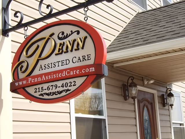 Close-up of a hanging sign for Penn Assisted Care outside a beige building with a window and a door with glass panels, along with two wall-mounted lantern-style lights.