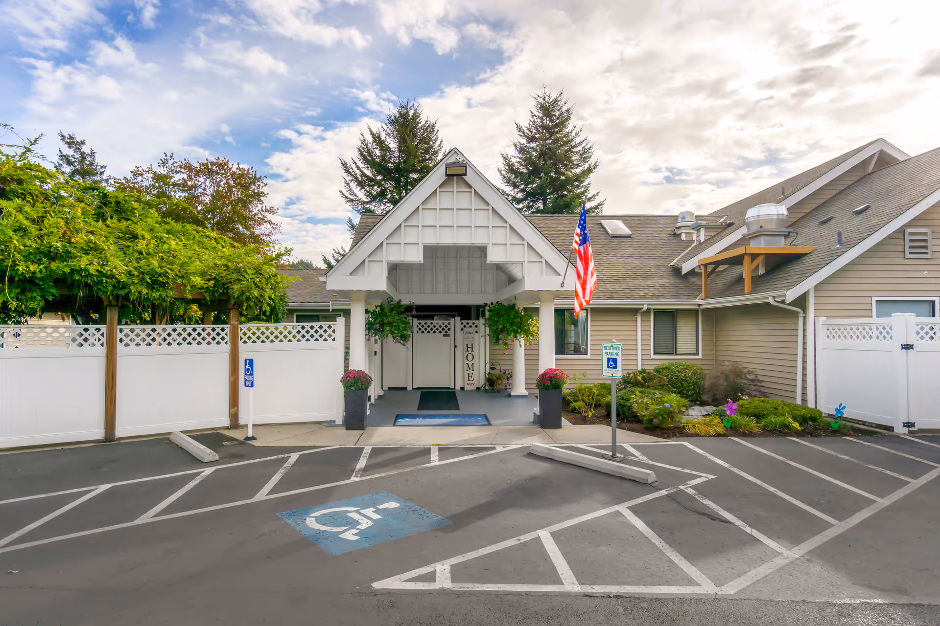 Entrance of a senior living facility with a covered porch, two large planters with flowers, an American flag, and designated handicapped parking spaces in front. The building is surrounded by greenery and a white fence under a partly cloudy sky.