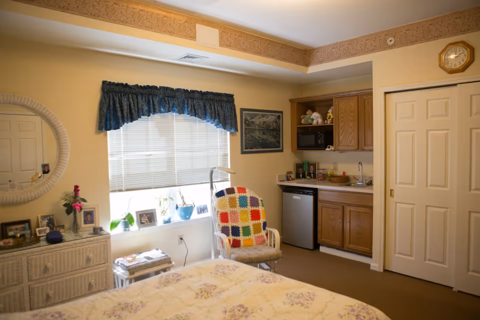 A cozy studio bedroom with a bed in the foreground, dresser and window, an armchair with a colorful afghan, and a small kitchenette area.