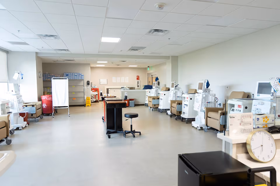 A bright and clean medical treatment room with multiple dialysis machines lined up along the right wall, each paired with a beige recliner chair. The room has a light-colored floor and ceiling with fluorescent lighting. There are medical carts, a clock, and various medical supplies visible throughout the space.