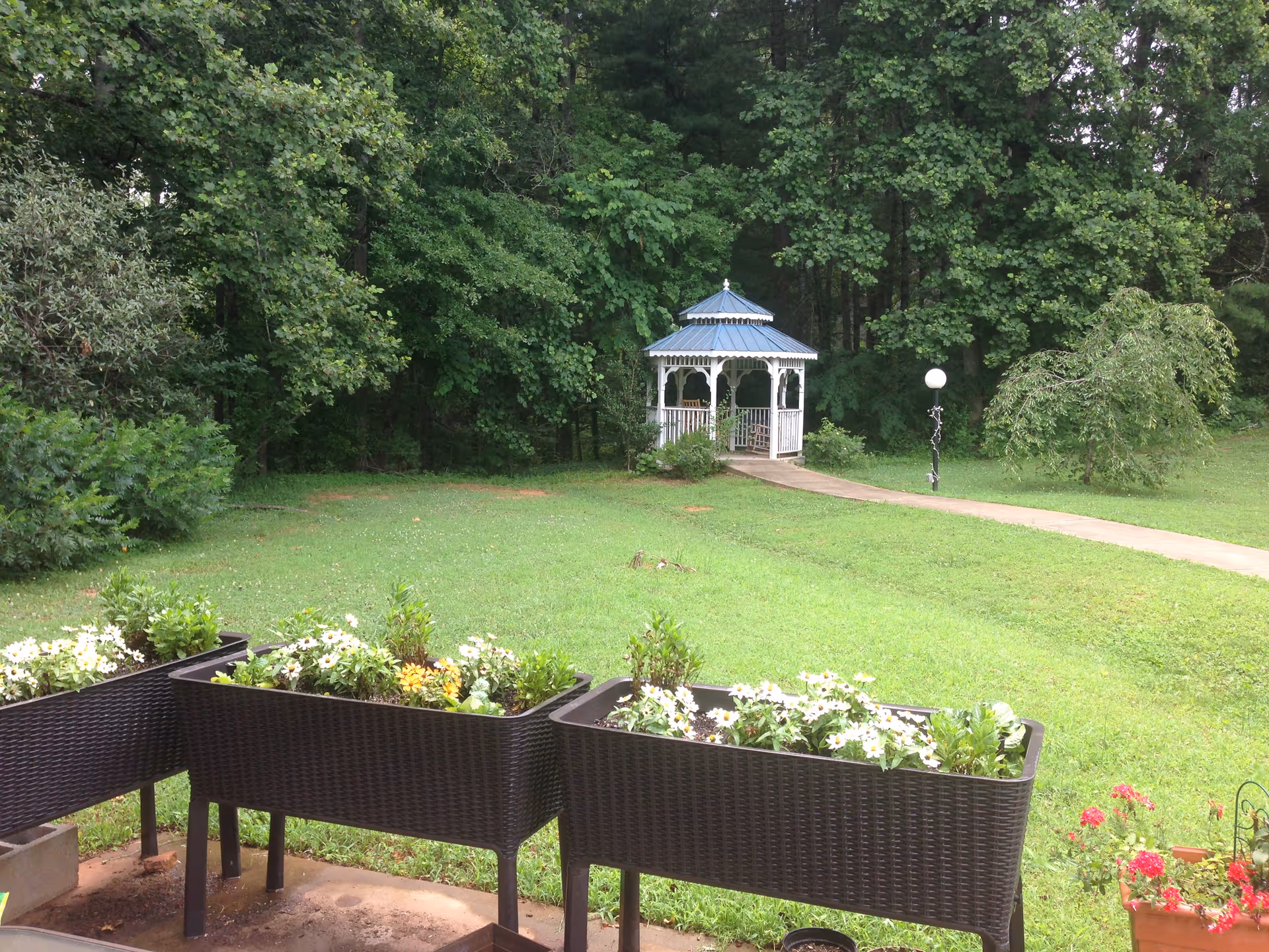 Green lawn with planter boxes in the foreground and a white gazebo along a path in front of a tree line.