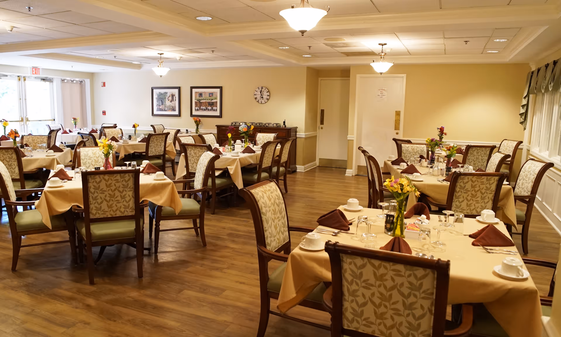 A well-lit dining room with multiple tables covered in beige tablecloths, each set with cups, glasses, silverware, and brown folded napkins. The chairs have wooden frames with patterned upholstery. Small flower vases decorate each table. The room has wooden flooring, beige walls, ceiling lights, and framed pictures on the wall.
