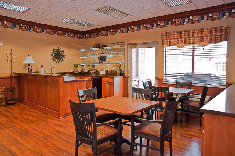 A cozy dining area with wooden tables and chairs, a small kitchen counter with a sink, glass shelves holding glassware, a window with blinds and a plaid valance, and warm wood flooring and cabinetry.