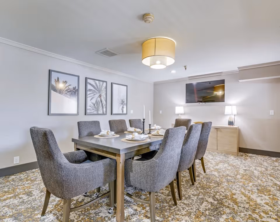 A dining room with a rectangular table set with place settings, surrounded by upholstered chairs beneath a ceiling light, with framed wall art and a wall-mounted TV.