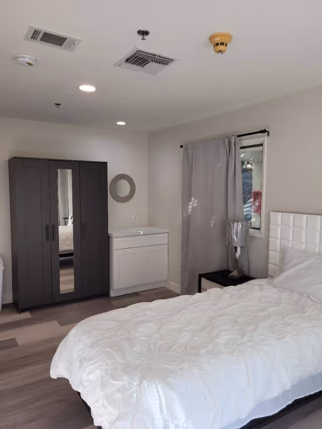 A bedroom with a white bed covered with a white quilt, a black nightstand with a silver lamp, a window with gray curtains, a white cabinet with a round mirror above it, and a dark wardrobe with a mirror on one door. The room has light-colored walls and wood flooring.
