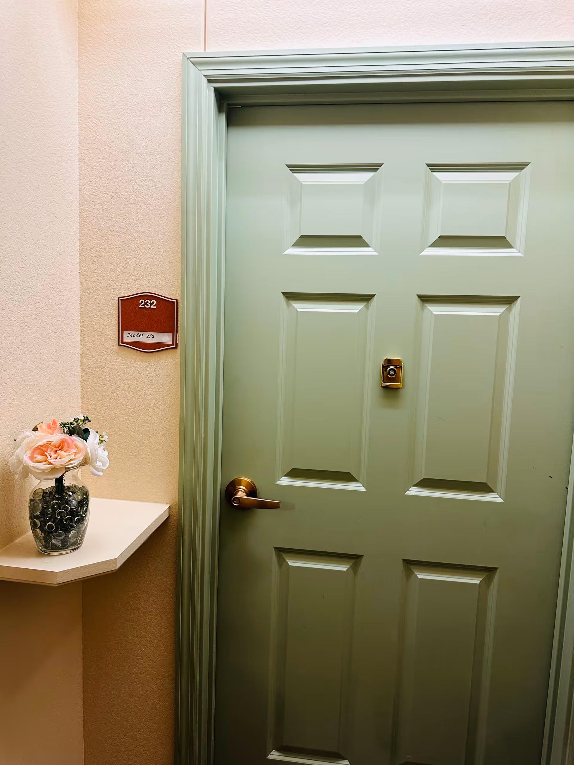 A closed green door with a brass handle and peephole in a hallway. To the left of the door is a small wall-mounted shelf holding a glass vase with pink and white flowers. A red sign next to the door reads '232 Model 2/2'.