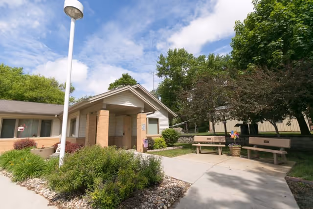 Exterior view of Careage Hills Rehabilitation And Healthcare facility showing a single-story building with a covered entrance, surrounded by greenery and trees. There are two benches and a potted plant with a colorful pinwheel on a paved area near the entrance under a partly cloudy sky.