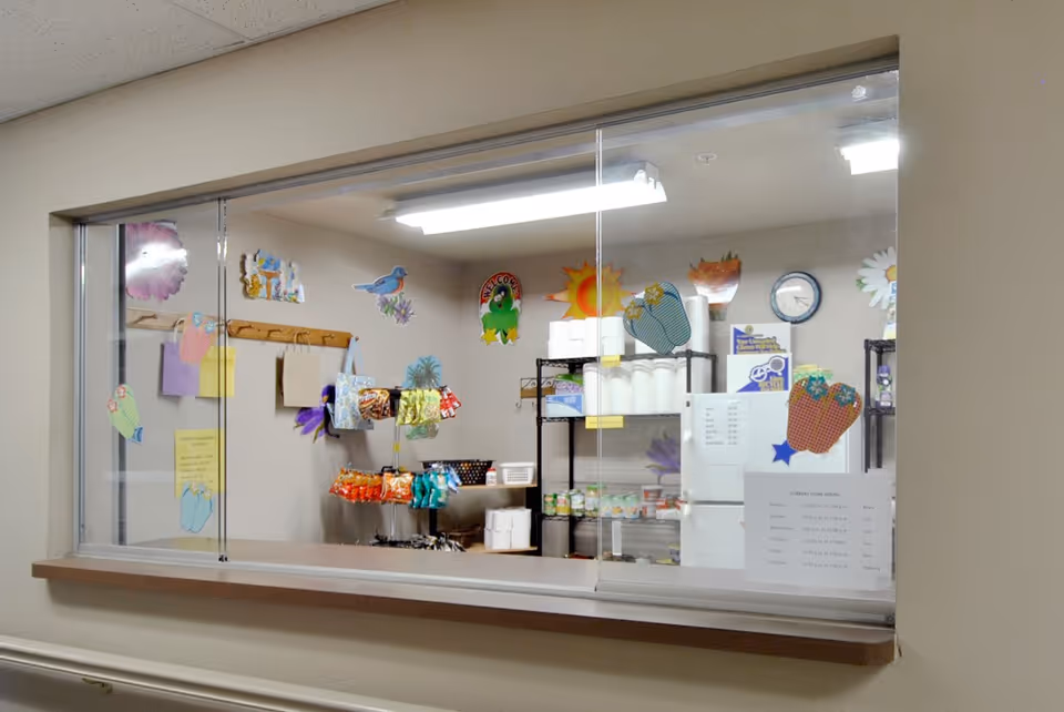 A service window looking into a small pantry with shelves, a refrigerator, snacks, and colorful paper decorations on the walls.