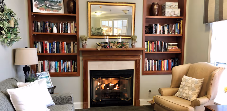 A cozy living room area featuring a lit fireplace with a wooden mantel decorated with candles and greenery. On either side of the fireplace are built-in wooden bookshelves filled with books and decorative items. To the left is a gray upholstered sofa with white pillows and a side table with a lamp and framed photo. To the right is a beige armchair with a patterned cushion, next to a window with a beige curtain.