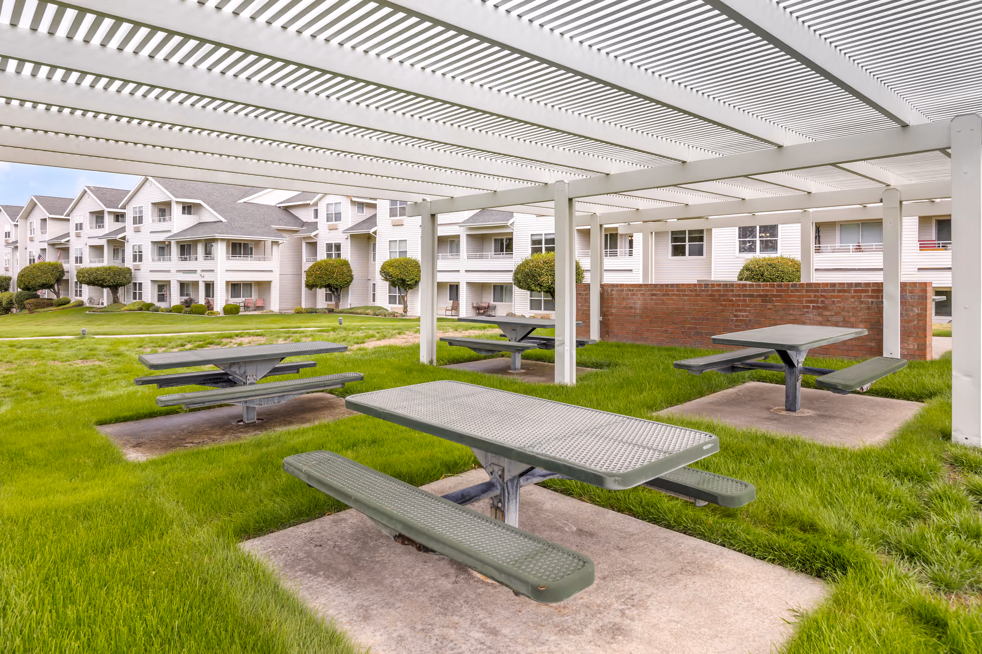 Outdoor covered picnic area with multiple green metal picnic tables on concrete pads surrounded by grass, with a multi-story residential building and trimmed bushes in the background.