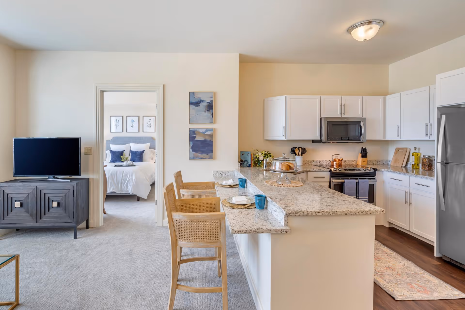 View of a modern open-concept living space featuring a kitchen with white cabinets, granite countertops, and stainless steel appliances. There is a kitchen island with three wooden bar stools and place settings. To the left, a doorway leads to a bedroom with a neatly made bed and framed artwork above the headboard. A TV sits on a dark wooden console against the wall near the doorway.