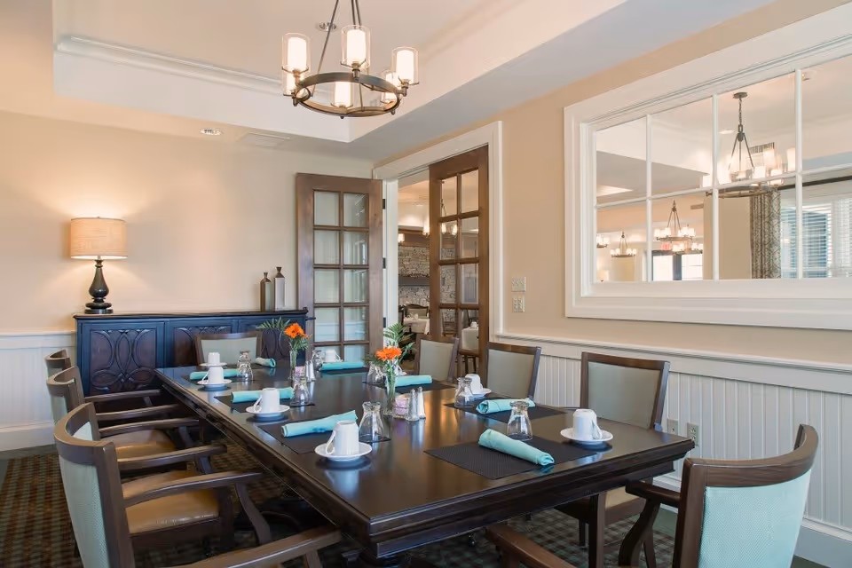 A dining room with a long dark wooden table set for eight people. Each place setting has a white cup and saucer, a glass turned upside down, and a turquoise cloth napkin. There are small vases with orange flowers on the table. The room has beige walls, a sideboard with a lamp and decorative items, and a large window with white trim looking into another room. A chandelier with multiple lights hangs above the table.