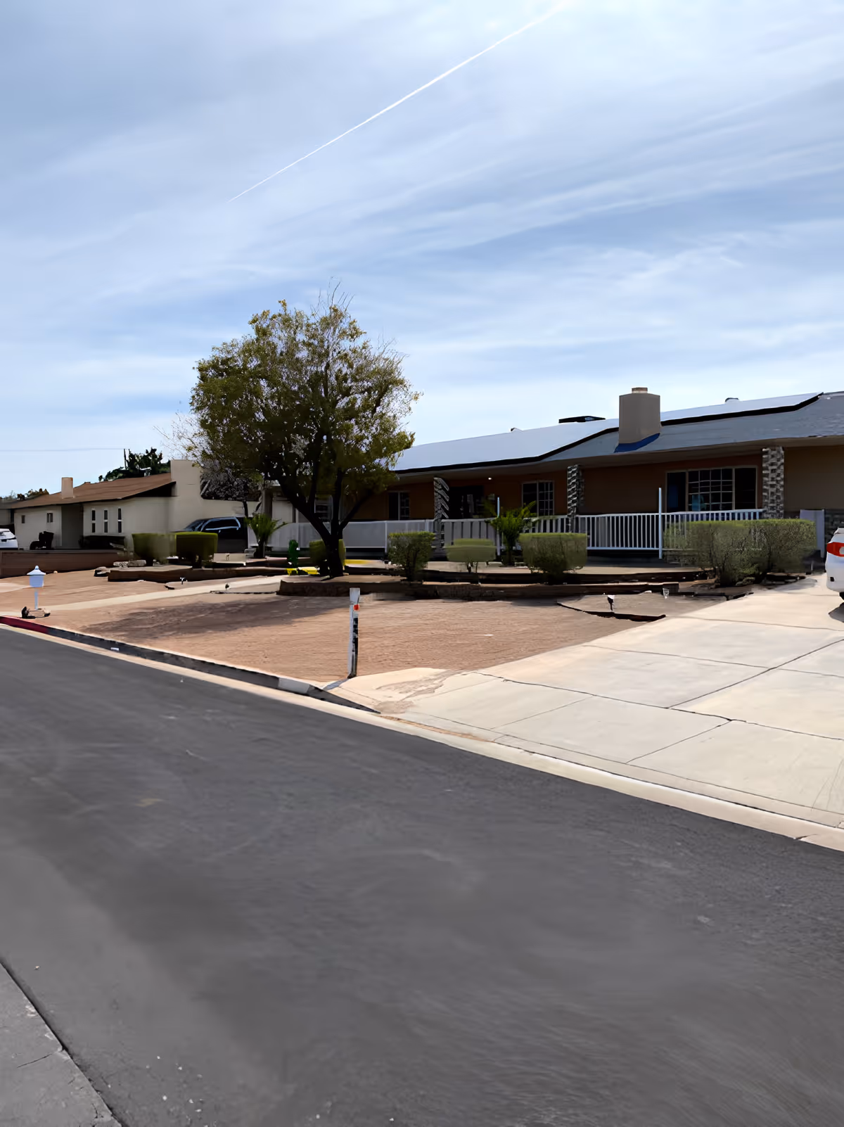 Exterior view of a single-story residential building with a porch, surrounded by trimmed bushes and a tree in a dry, landscaped yard. The sky is partly cloudy with a visible contrail.