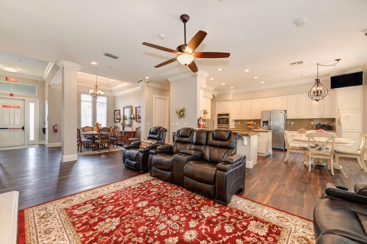 Spacious senior living facility common area featuring a seating area with black leather recliners on a red patterned rug, a dining area with a wooden table and chairs, and a kitchen with white cabinetry, stainless steel appliances, and a small round dining table. The room has hardwood floors, a ceiling fan, and recessed lighting.