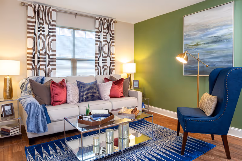 A cozy living room with a beige sofa adorned with multiple colorful pillows, a blue throw blanket, and two side tables with lamps. A glass coffee table with decorative items sits on a blue patterned rug. A blue armchair with a pillow and a floor lamp are positioned near a green accent wall with a large abstract painting.