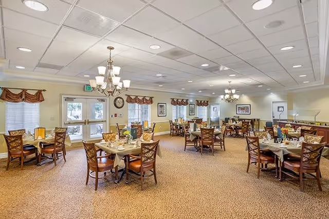 A spacious dining room with multiple round tables set for meals, each surrounded by wooden chairs with cushioned seats. The room has beige walls, carpeted floors, and several windows with valance curtains. Two chandeliers hang from the ceiling, and framed artwork decorates the walls. Double doors with exit signs are visible at the far end.