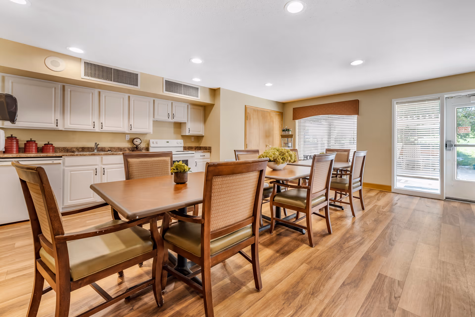 A bright dining room with wooden floors and a long wooden table surrounded by cushioned chairs. The room features white kitchen cabinets, a stove, and a countertop with decorative items. Large windows with blinds and a glass door allow natural light to enter the space.