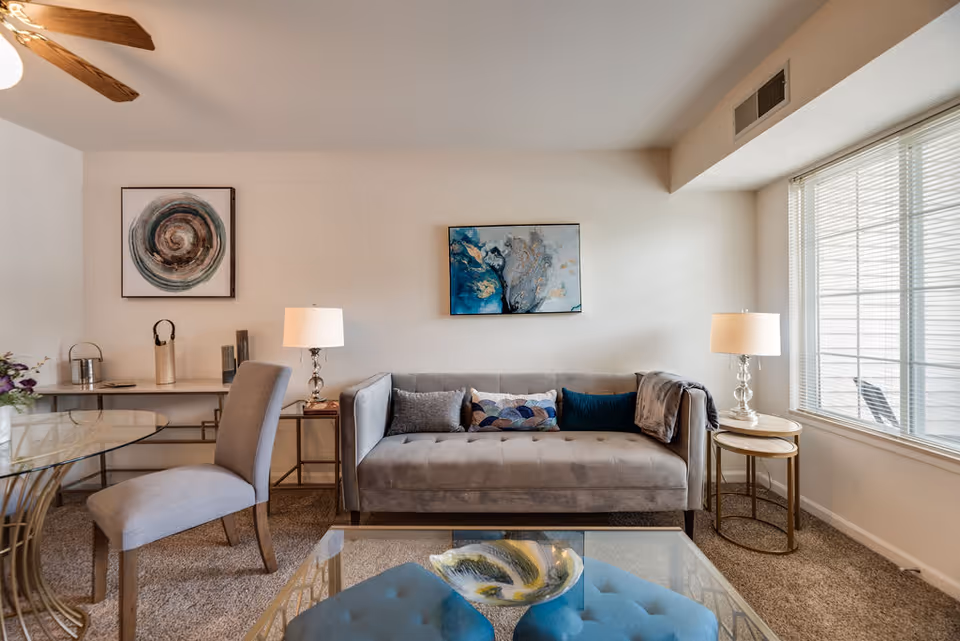 A cozy living room with a gray tufted sofa adorned with decorative pillows and a throw blanket. Two side tables with lamps flank the sofa. A glass coffee table with a decorative bowl sits in front of the sofa. A gray upholstered chair is positioned near a glass dining table with gold legs. The room features two abstract paintings on the walls, a ceiling fan with wooden blades, and a large window with blinds letting in natural light.