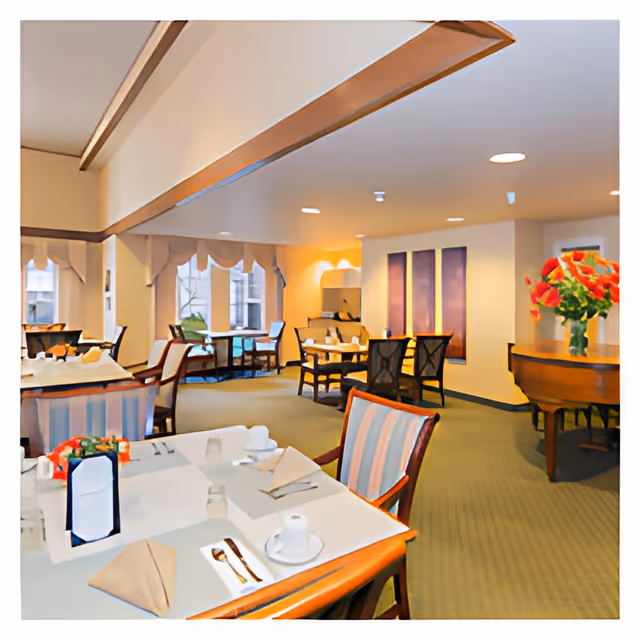A dining room in a senior living facility with several tables set with napkins, cups, and utensils. The room has carpeted floors, striped upholstered chairs, large windows letting in natural light, and a piano with a vase of red flowers on top.