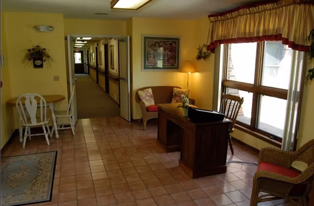 Interior view of a senior living facility hallway with a small seating area. The seating area includes a wooden desk with a laptop, wicker chairs with cushions, a small round table with two white chairs, a floor lamp, and framed artwork on the wall. Large windows with curtains let in natural light, and the floor is tiled.