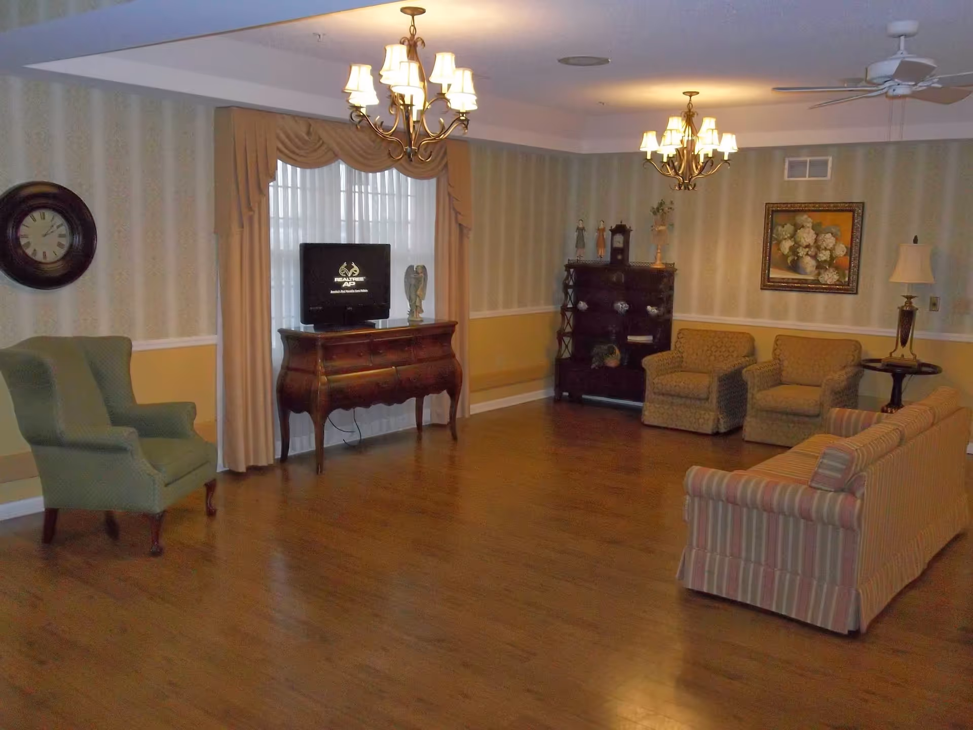 A cozy living room with wooden flooring, featuring a striped sofa, two patterned armchairs, and a green armchair. There is a wooden cabinet with a TV on top, a wall clock, a painting of flowers, two chandeliers, and a ceiling fan. The walls are decorated with striped wallpaper and a chair rail with a yellow lower half.