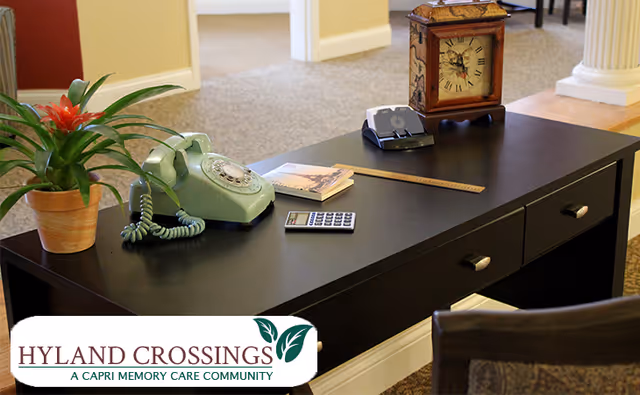 A black desk with various items on it including a green rotary phone, a potted plant with red flowers, a wooden clock, a calculator, a ruler, and some papers. The desk is in a carpeted room with beige walls and white trim, with doorways visible in the background.