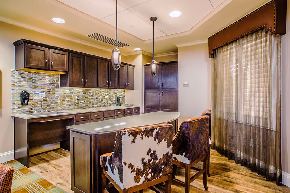 A modern kitchen area with dark wooden cabinets, a mosaic tile backsplash, and a light-colored countertop. There is a kitchen island with two high-back chairs upholstered in a brown and white cowhide pattern. Two pendant lights hang above the island, and a window with sheer brown curtains is on the right side.