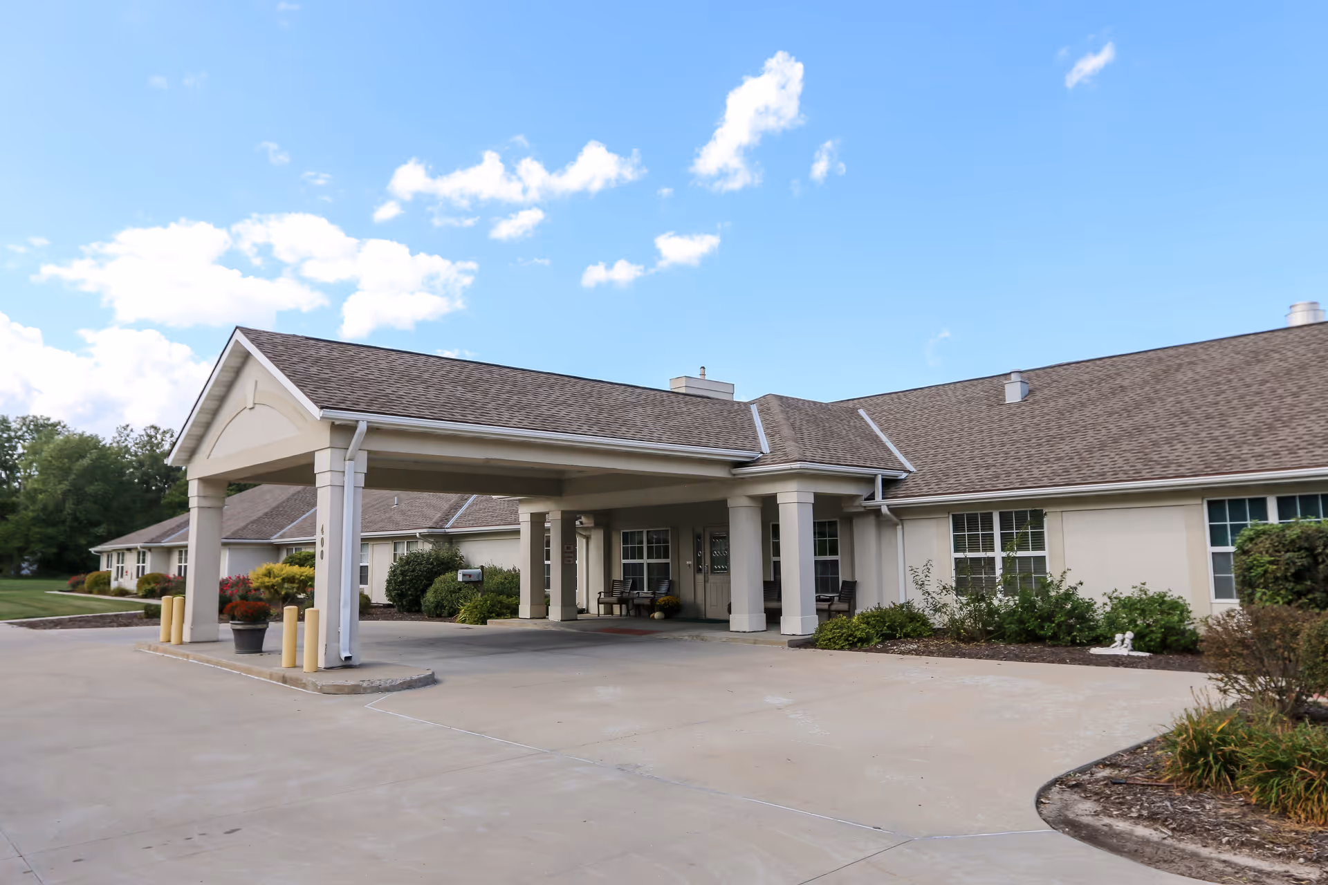 Single-story senior living facility entrance with a covered porte-cochère, columns, and landscaped grounds under a blue sky.