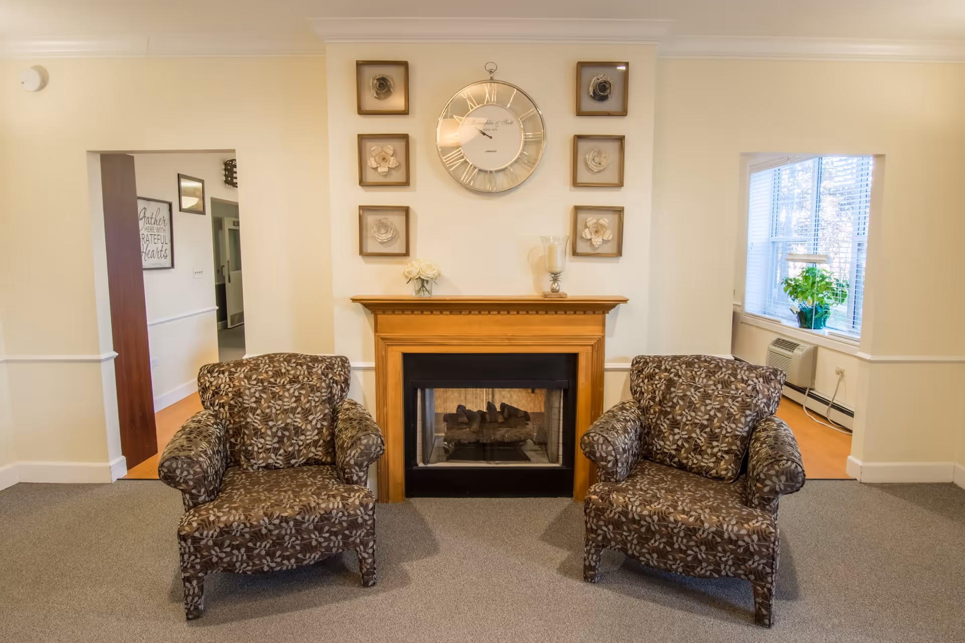 A cozy living room area with two patterned armchairs facing a wooden fireplace. Above the fireplace is a large round clock surrounded by six framed floral artworks. To the right, there is a window with blinds and a green potted plant on the windowsill. The room has beige walls and carpeted flooring.