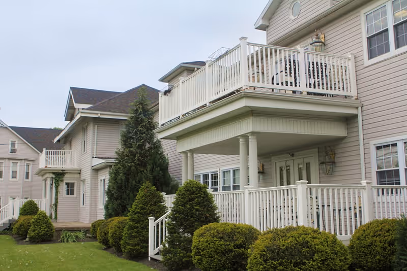 Exterior view of a multi-story senior living building with white railings, balconies, entrances and neatly trimmed shrubs.