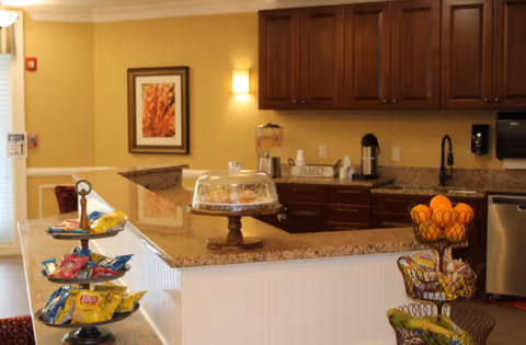Open serving kitchen area with a granite countertop, snack displays, wooden cabinets, and a cake under a glass dome.