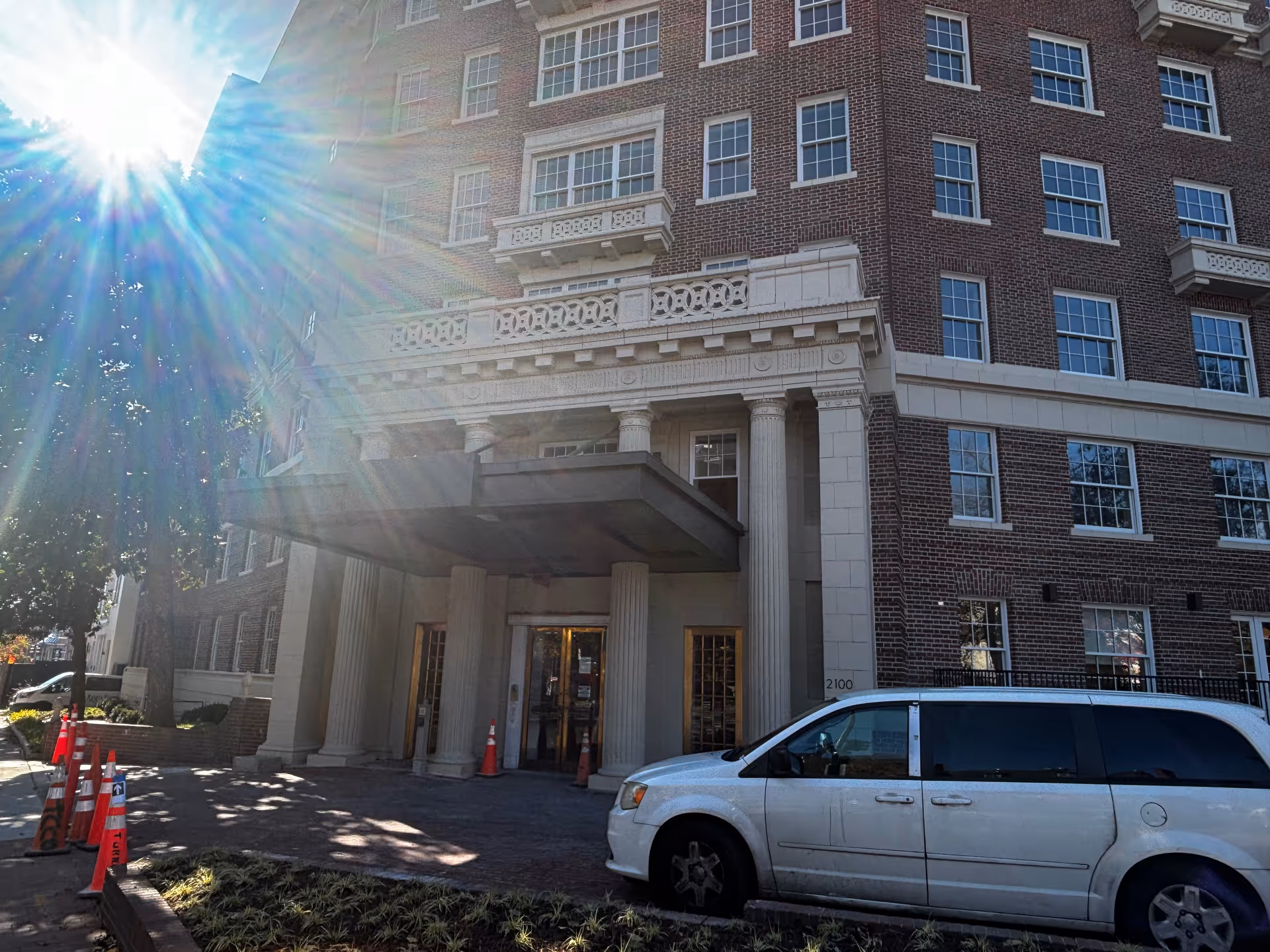 Front entrance of a multi-story brick building with classical columns and a covered porte-cochère, a white van parked in front and a bright sun flare in the upper left.
