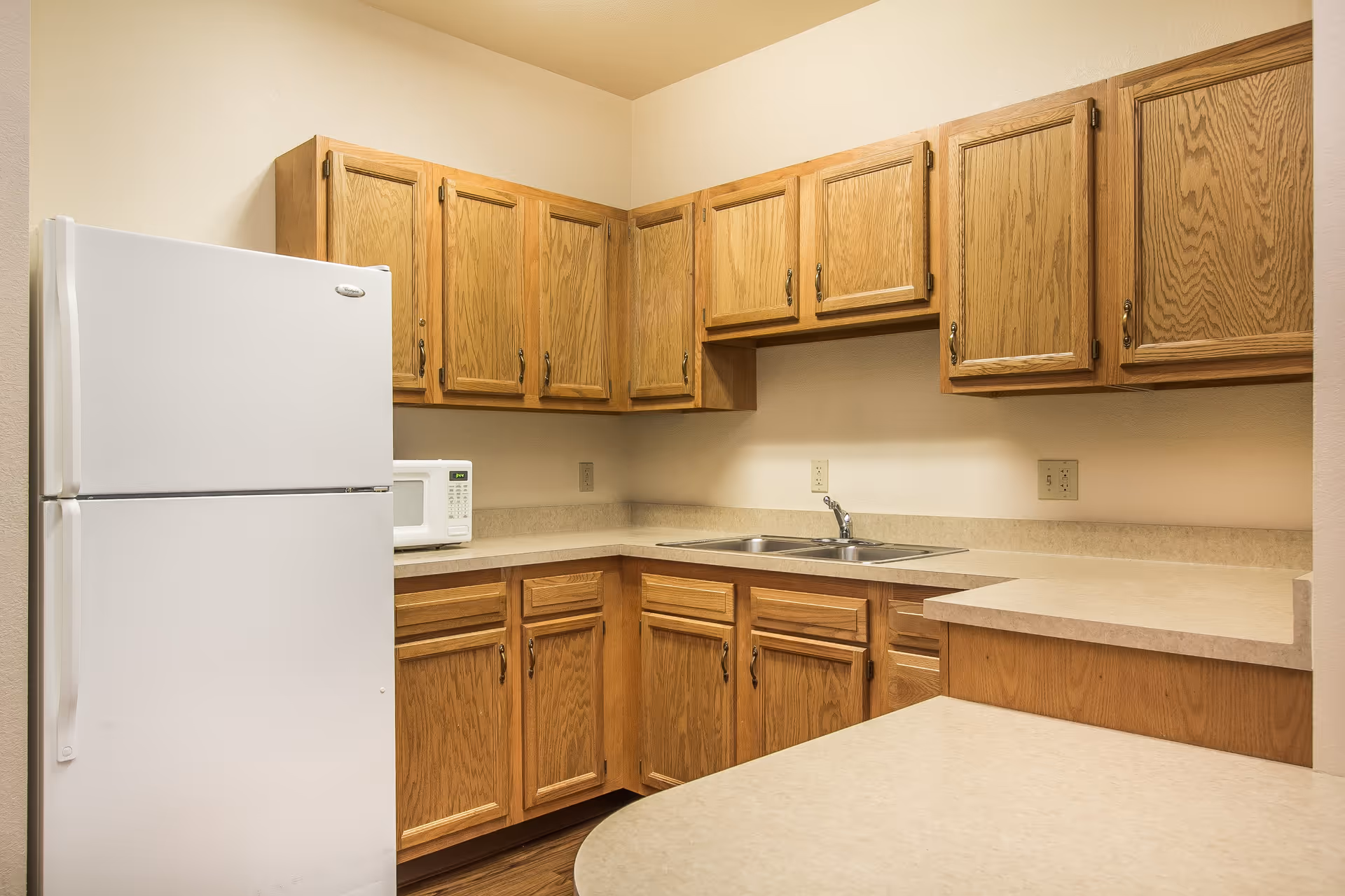 A kitchen with wooden cabinets, a white refrigerator, a microwave, a double sink, and beige countertops.