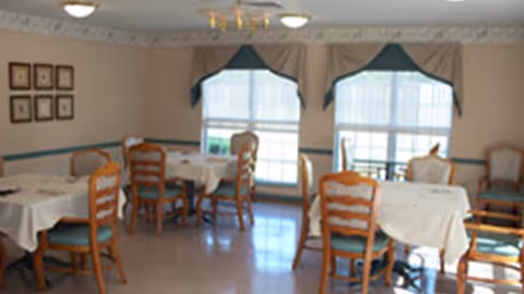 A dining room with several tables covered with white tablecloths and wooden chairs with green cushions. The room has large windows with beige curtains and valances, and framed pictures on one wall. The floor is light-colored and the room is well-lit with ceiling lights.