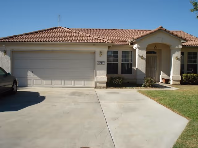 Front exterior view of a single-story house with a tiled roof, a two-car garage with a closed door, a driveway, a small front porch with an arched entryway, and some shrubs near the windows. The house number 2226 is visible on the wall next to the garage.