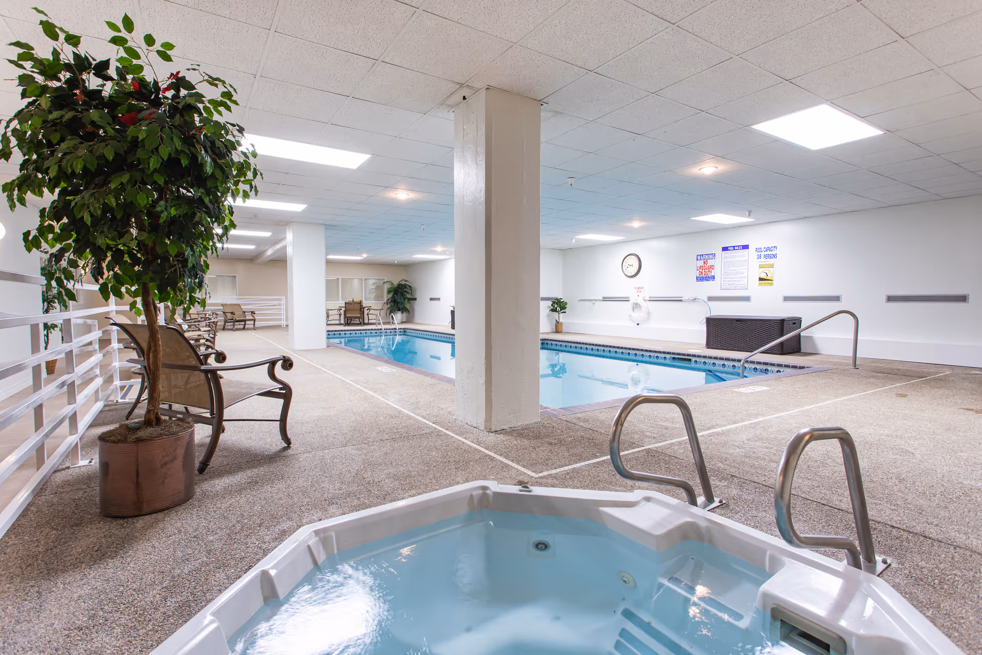 Indoor swimming pool area with a hot tub in the foreground, metal handrails, potted plants, and several chairs along the side. The space has a white ceiling with recessed lighting and white walls with safety signs and a clock.