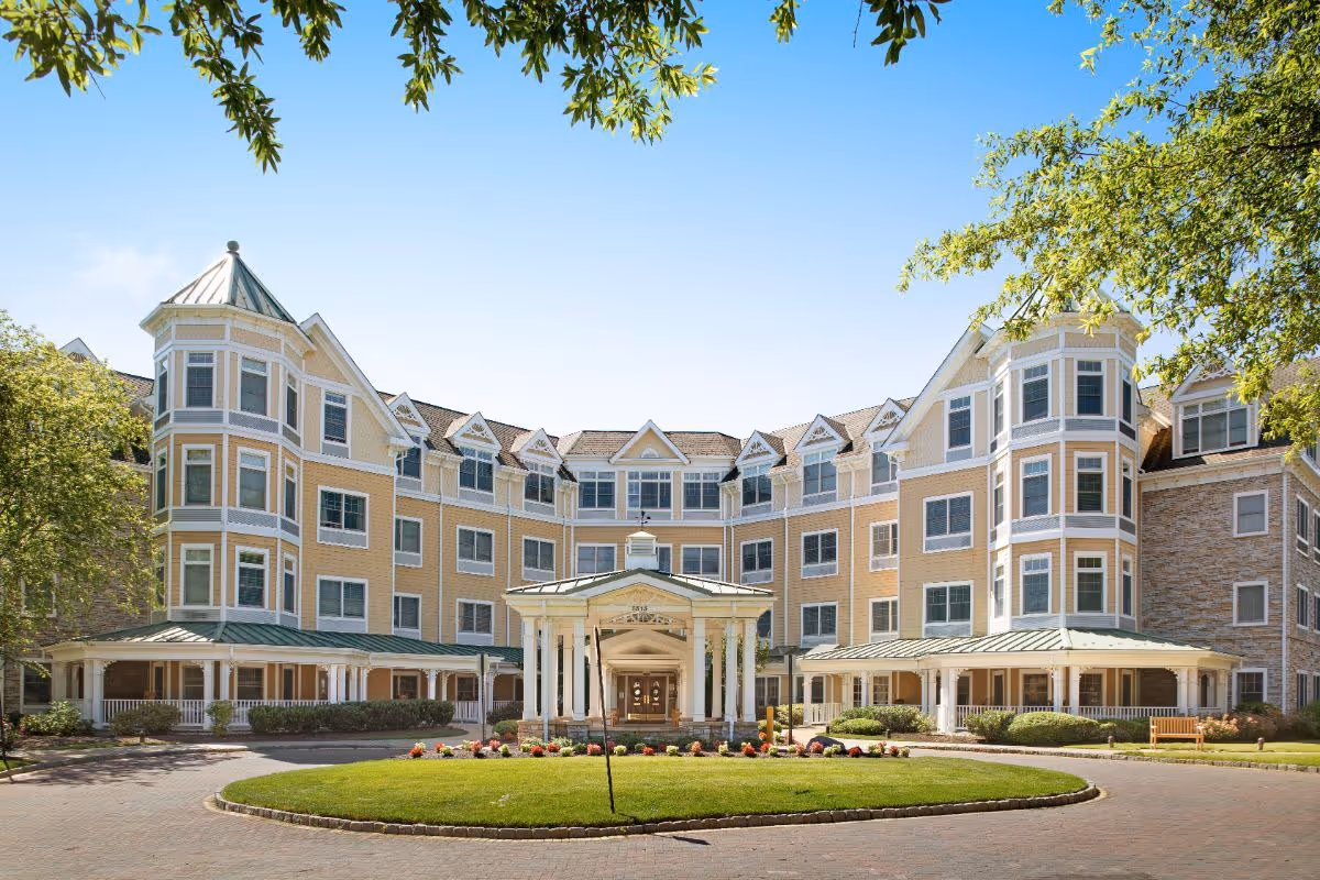 Front exterior view of a large, multi-story senior living facility building with a circular driveway and a covered entrance. The building features multiple windows, light yellow siding with white trim, and a green metal roof. There are trees and landscaping around the driveway and building.