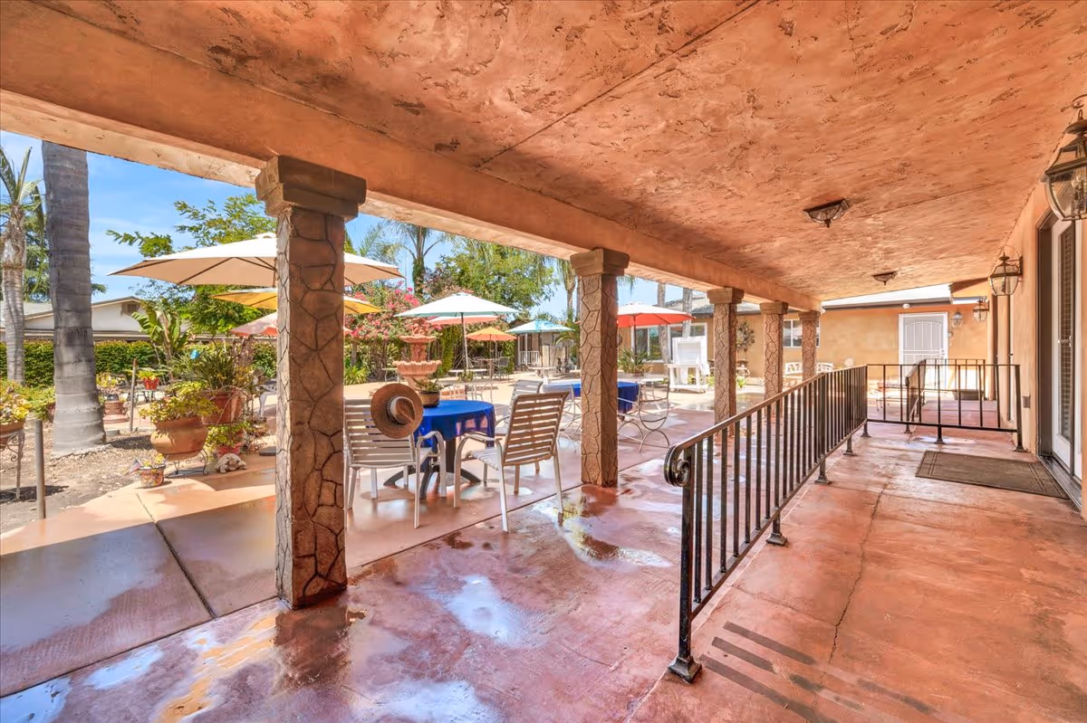 Covered patio area with stone pillars and a railing, overlooking an outdoor seating area with tables covered in blue tablecloths and chairs. Several umbrellas provide shade, and there are potted plants and palm trees in the background under a clear blue sky.