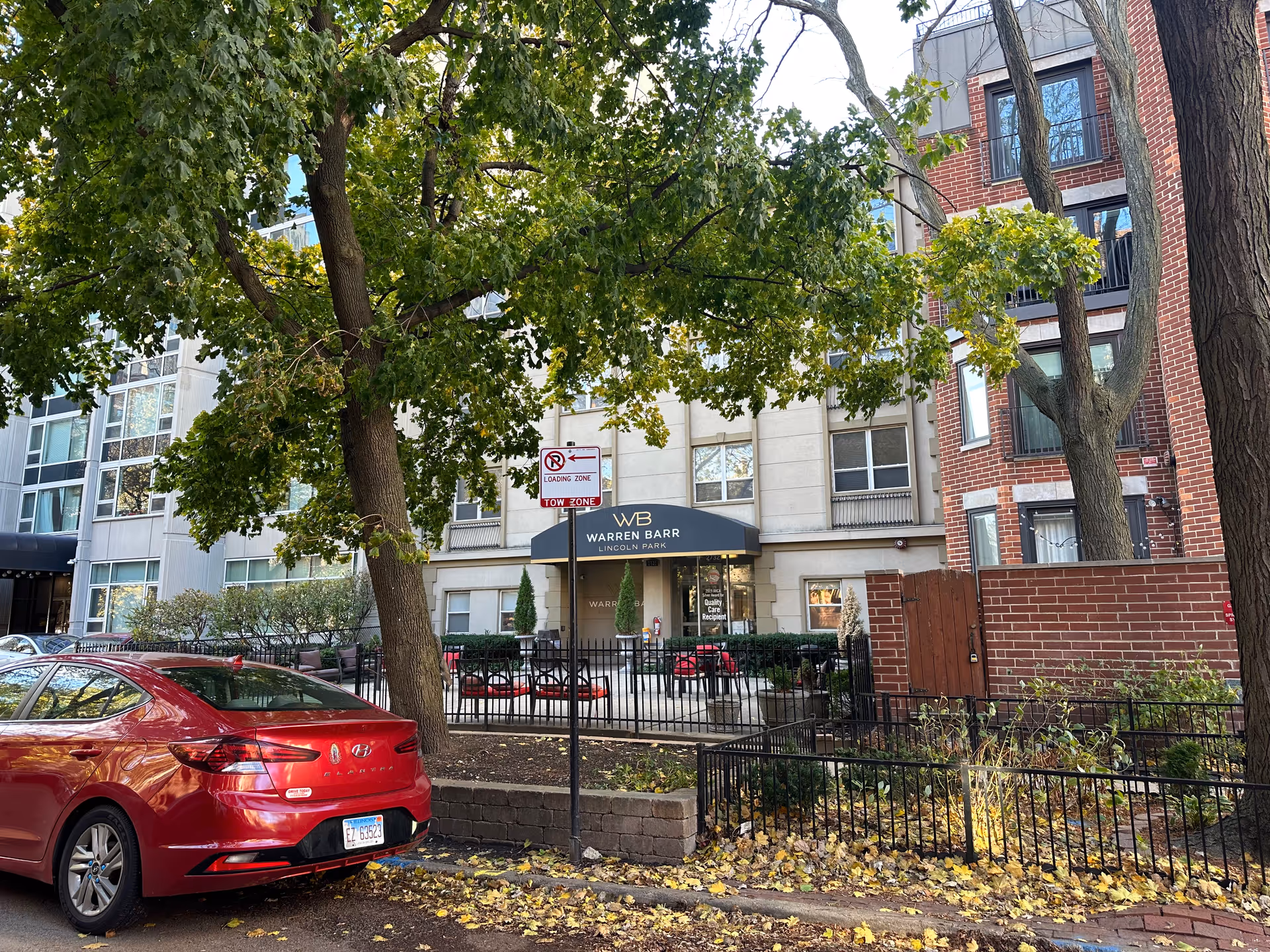 Exterior view of Warren Barr Lincoln Park facility showing the entrance with a black awning displaying the facility name. There are trees with green leaves, a red car parked on the street, and a fenced patio area with outdoor seating. The building is multi-story with a mix of beige and red brick walls.
