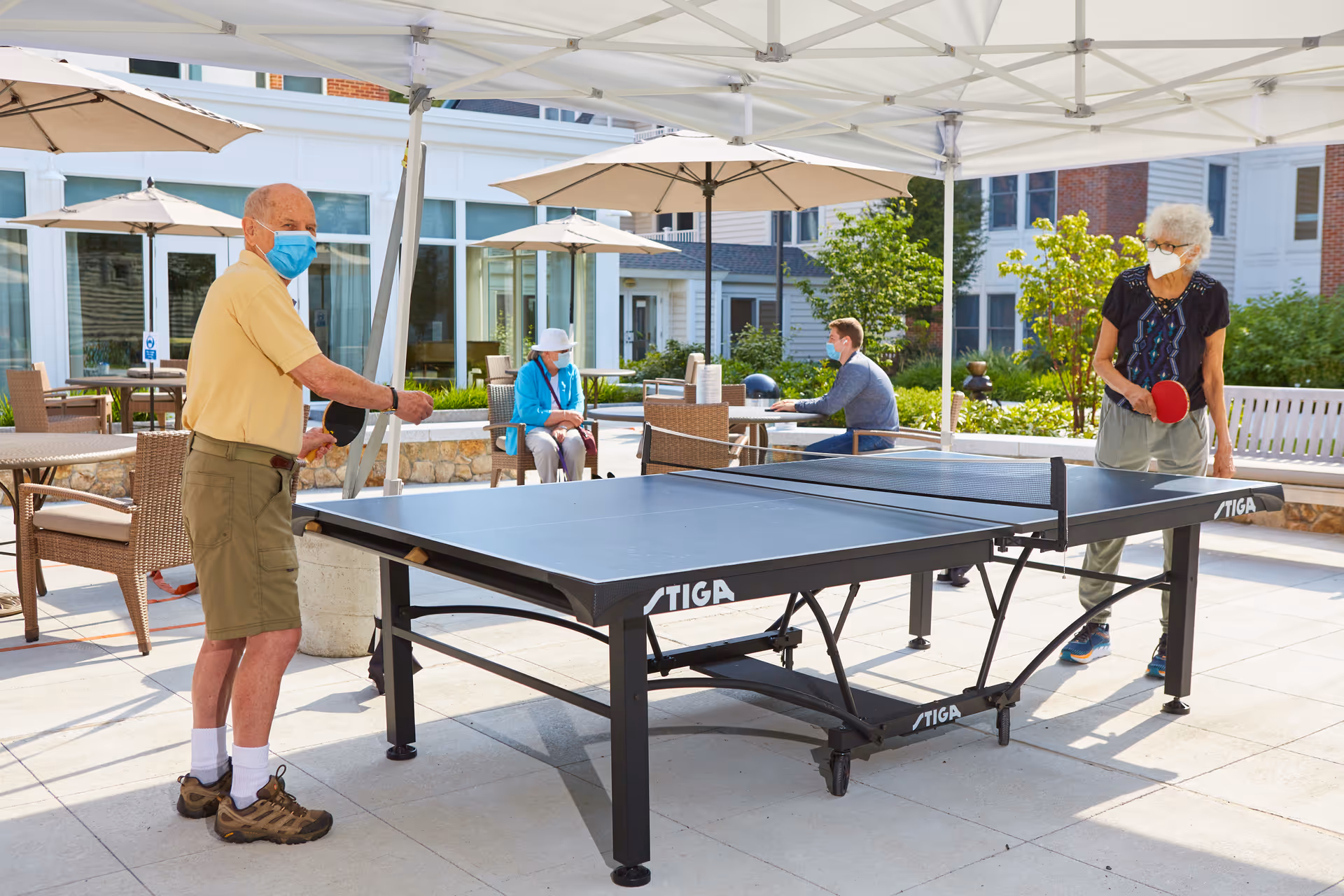 Two masked seniors play table tennis under a canopy on an outdoor patio while others sit nearby.