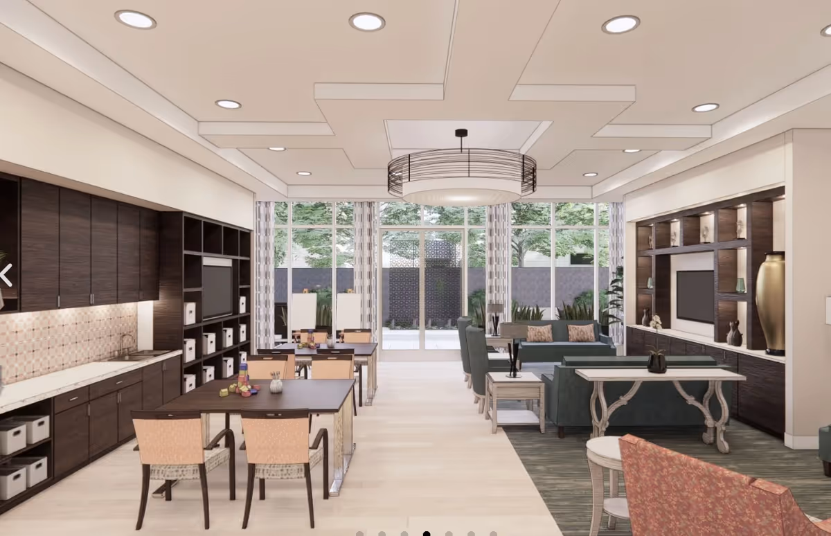 Bright communal living and dining area with seating, tables, a TV wall, and a kitchenette with dark cabinets in front of large windows.